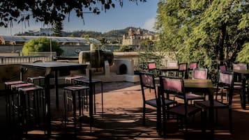 Un patio al aire libre con mesas y sillas, un muro de piedra y vistas a un paisaje urbano con edificios y una colina al fondo.