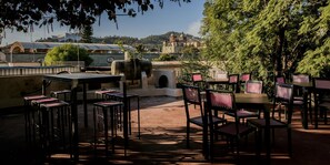 An outdoor patio with tables and chairs, a stone wall, and a view of a cityscape with buildings and a hill in the background.