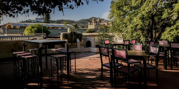 An outdoor patio with tables and chairs, a stone wall, and a view of a cityscape with buildings and a hill in the background.