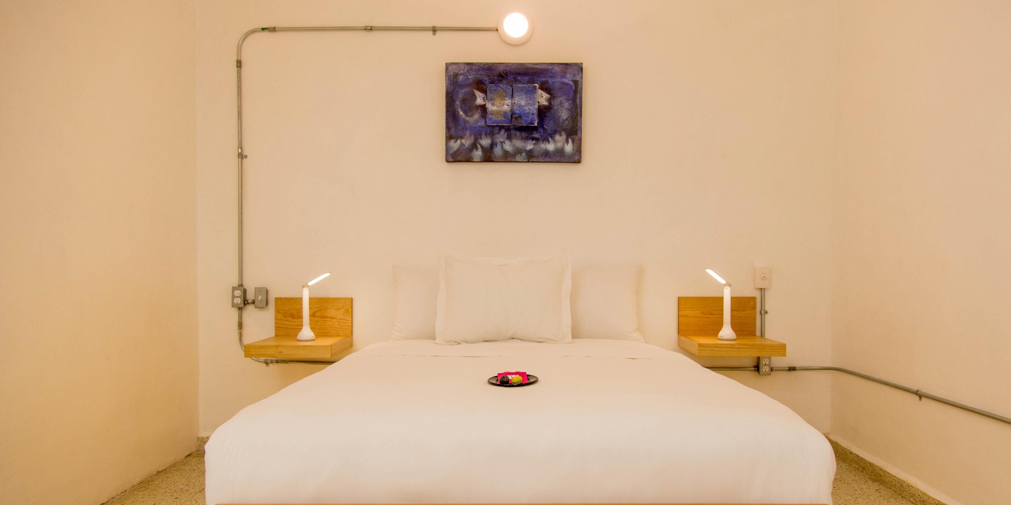 Una cama bien hecha con ropa blanca, dos mesitas de noche con lámparas, un cuadro en la pared y una lámpara colgante sobre la cama.