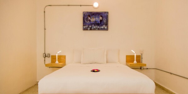 A neatly made bed with white bedding, two bedside tables with lamps, a wall-mounted painting, and a single light fixture above the bed.