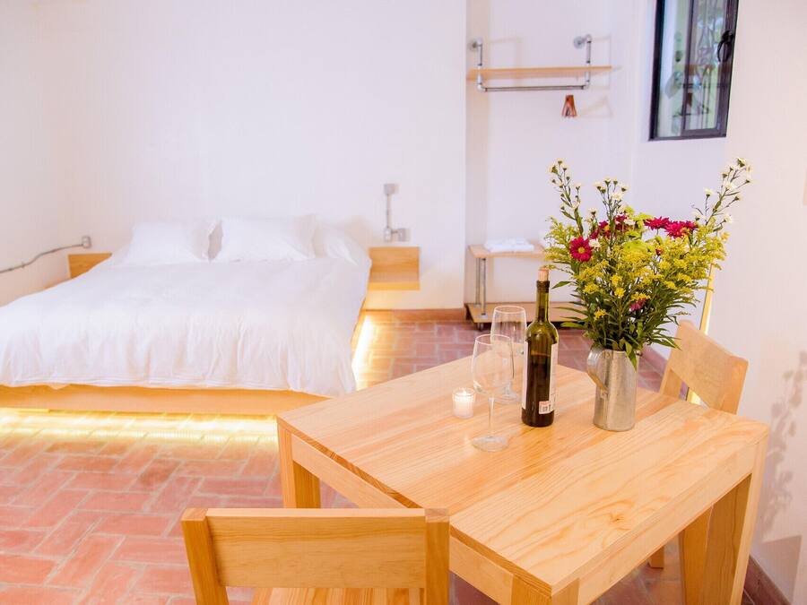 Una habitación de hotel con cama, una mesa de madera con sillas, un jarrón de flores y una botella de vino.