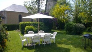 Un jardin avec une table et des chaises sous un parasol blanc, entouré de verdure et avec un bâtiment en arrière-plan.