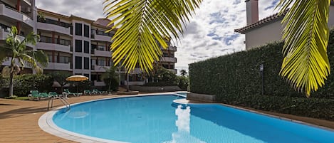 Una piscina con borde de baldosas, rodeada de palmeras y un edificio con varios balcones.