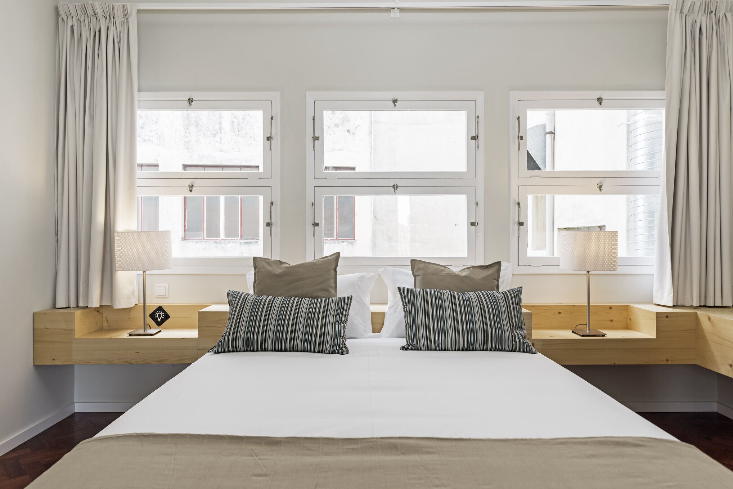 A bedroom with a large bed, two bedside tables, and three windows with curtains.