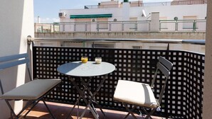 Ein Balkon mit Tisch und zwei Stühlen, mit Blick auf ein Gebäude mit grüner Markise und einer Umzäunung.