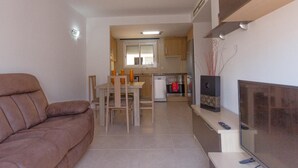 Ein Wohnzimmer mit braunem Sofa, einem Esstisch mit Stühlen, einer Küchenzeile mit Schränken und Geräten sowie einem Fernseher auf einem Schrank.