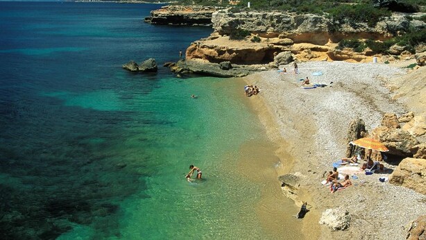 Ein Strand mit klarem, türkisblauem Wasser, felsigem Gelände und sonnenbadenden Menschen.