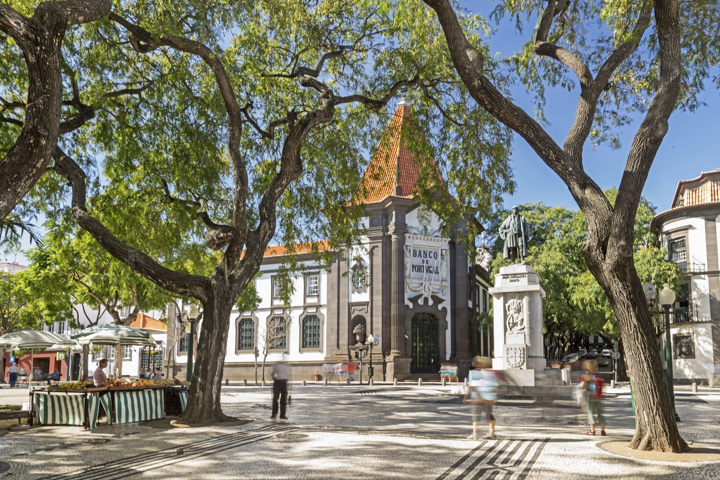 A plaza with a central monument, surrounded by trees and a building with a tiled roof.