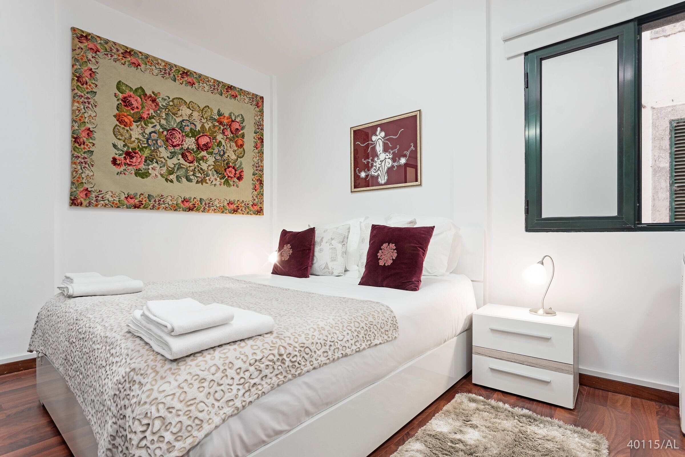 Une chambre à coucher comprenant un lit, une table de chevet, une lampe, un grand tableau représentant des motifs floraux et une œuvre d’art plus petite, encadrée.