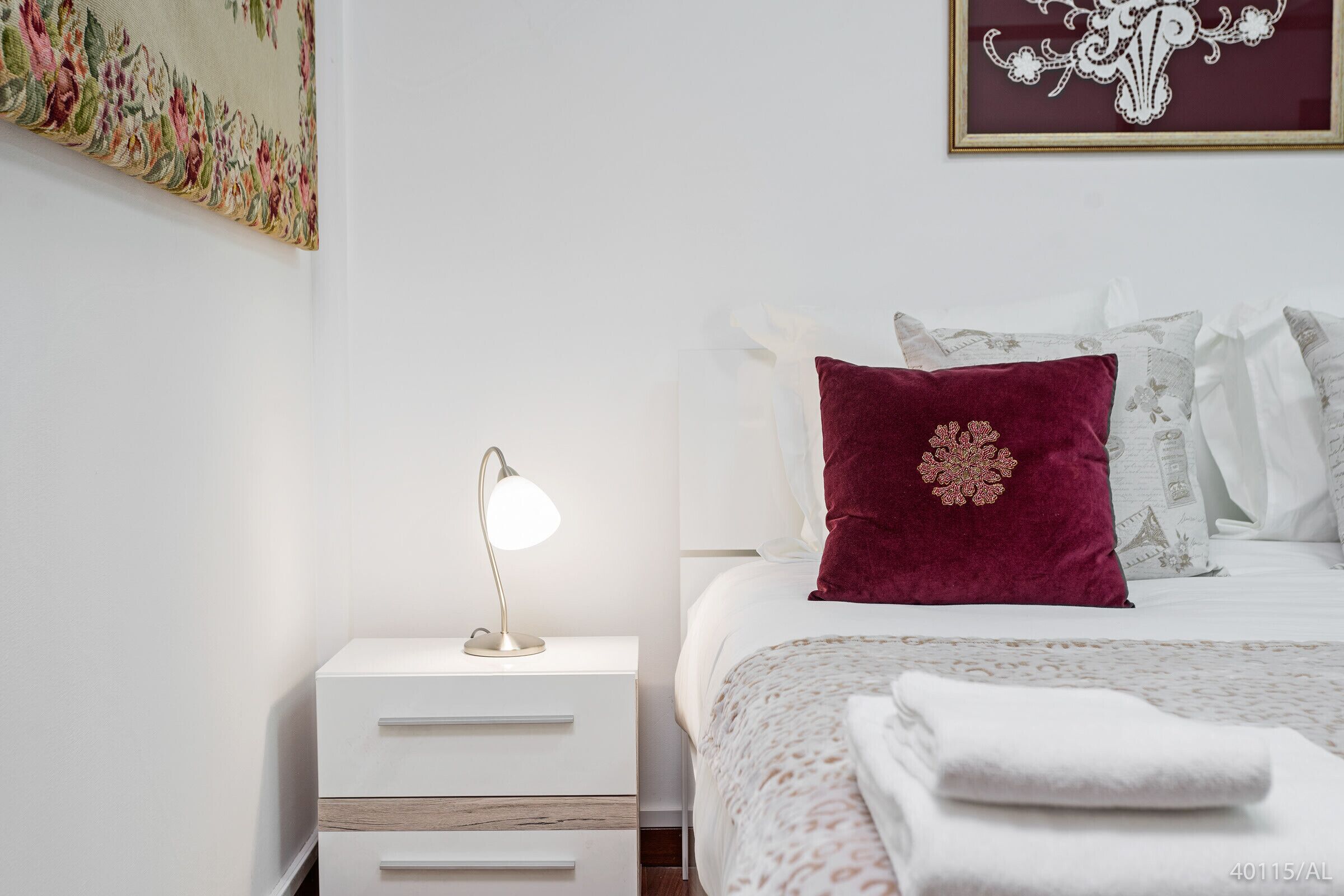 A neatly made bed with a patterned bedspread and a single decorative pillow. A bedside lamp is on a white nightstand with drawers.