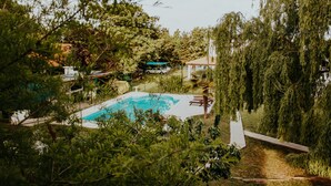 Une piscine entourée d’une végétation luxuriante, un bâtiment blanc et un véhicule garé.