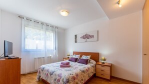 A bedroom with a bed, a desk, a nightstand, a lamp, a painting, and a window with curtains.