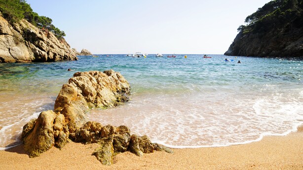 A beach with clear water, rocky formations, and boats in the distance.