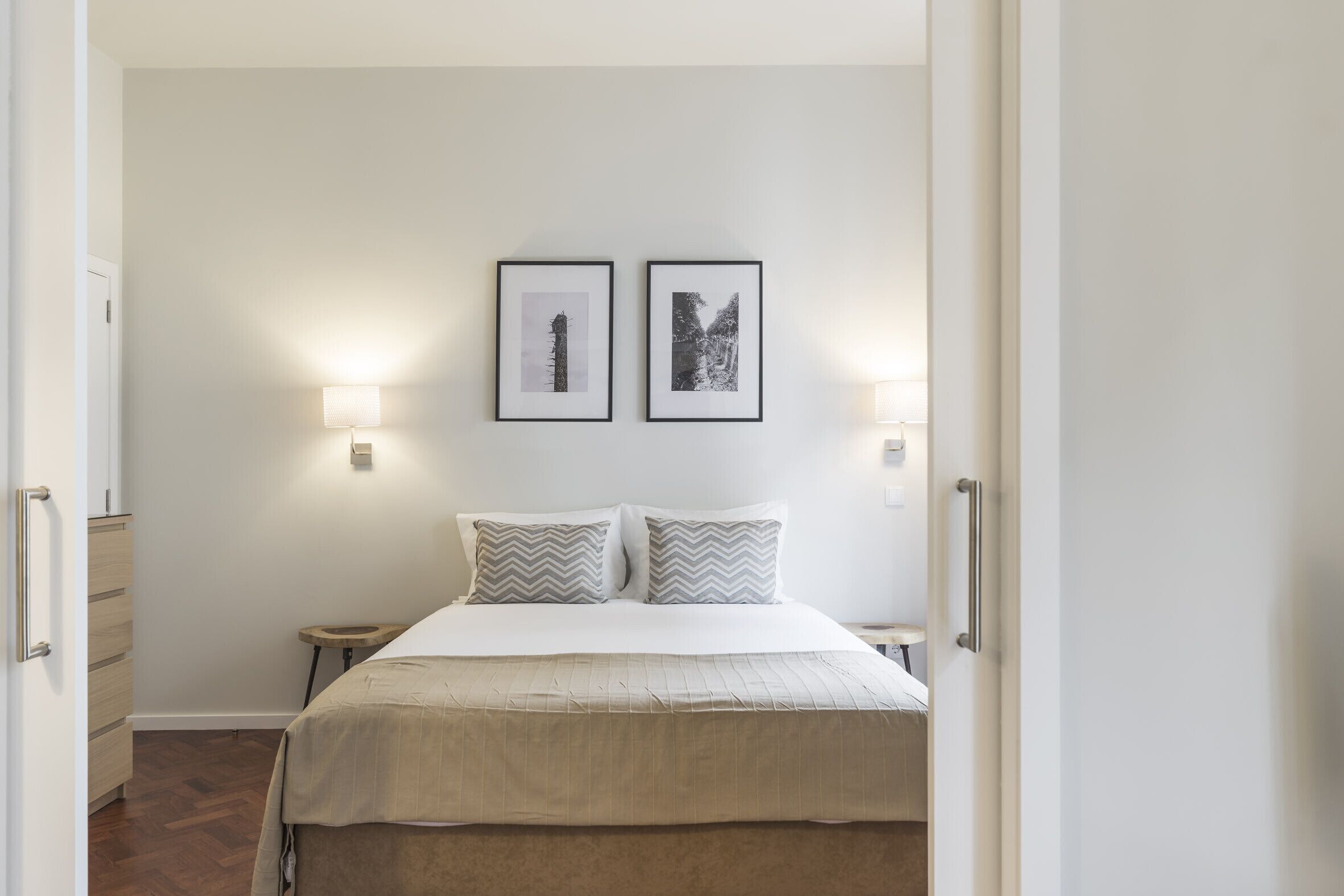 A bedroom with a bed, two pillows, a nightstand, a wall-mounted lamp, and two framed pictures.
