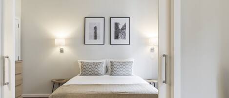 A bedroom with a bed, two pillows, a nightstand, a wall-mounted lamp, and two framed pictures.