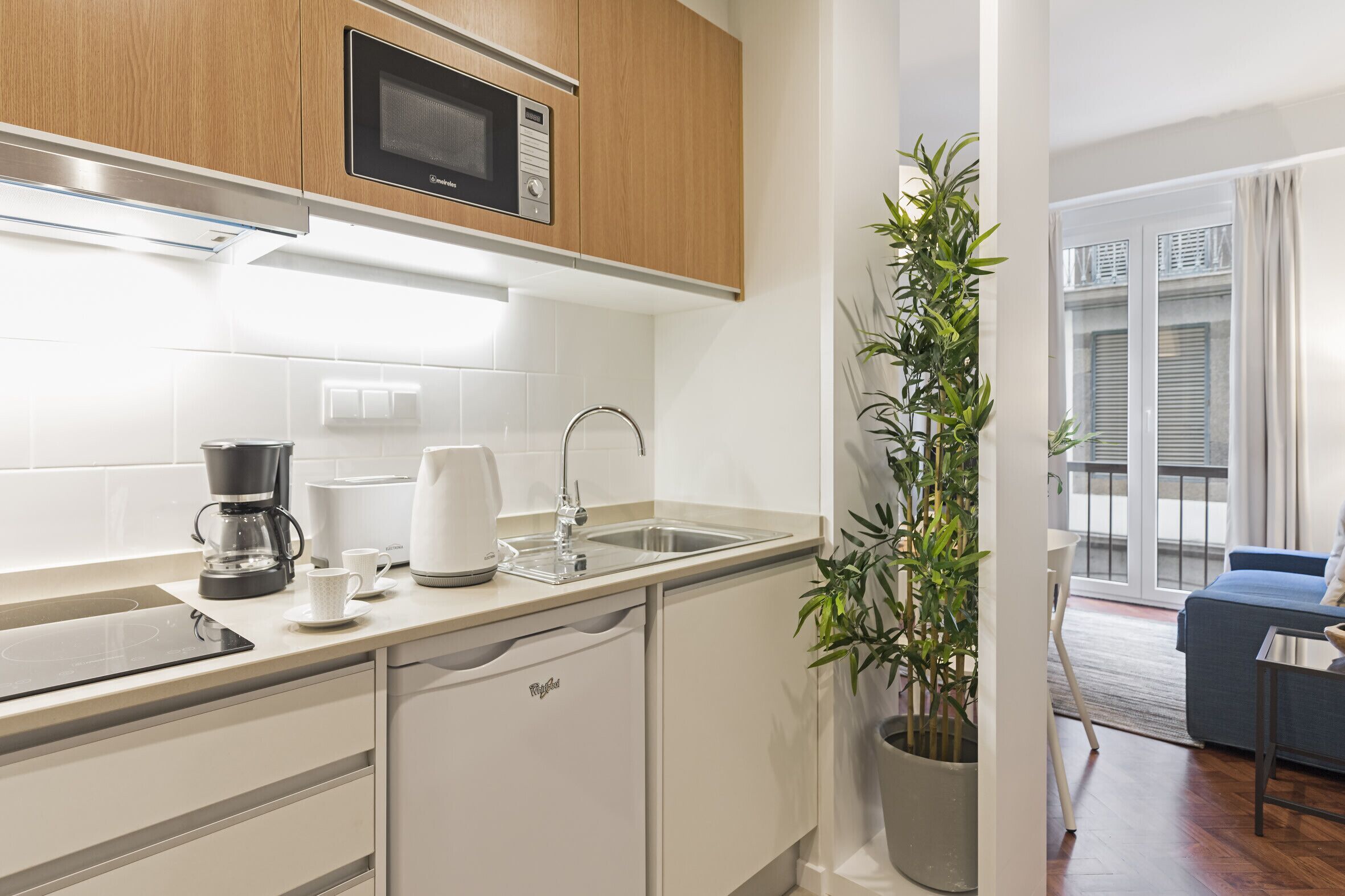 A modern kitchen with a microwave, coffee maker, and sink. There is a potted plant and a view of a balcony with a blue sofa.