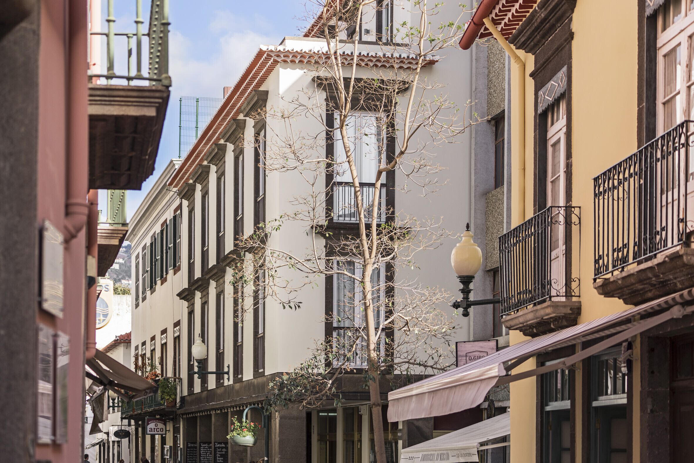 Uma rua estreita ladeada por edifícios altos, alguns com sacadas e toldos.