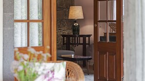 A room with a wooden door, a table with a lamp, a wicker chair, and a view through a window. - Casa do Lanço - Tinta Negra, wine and laurissilva. (São Vicente)