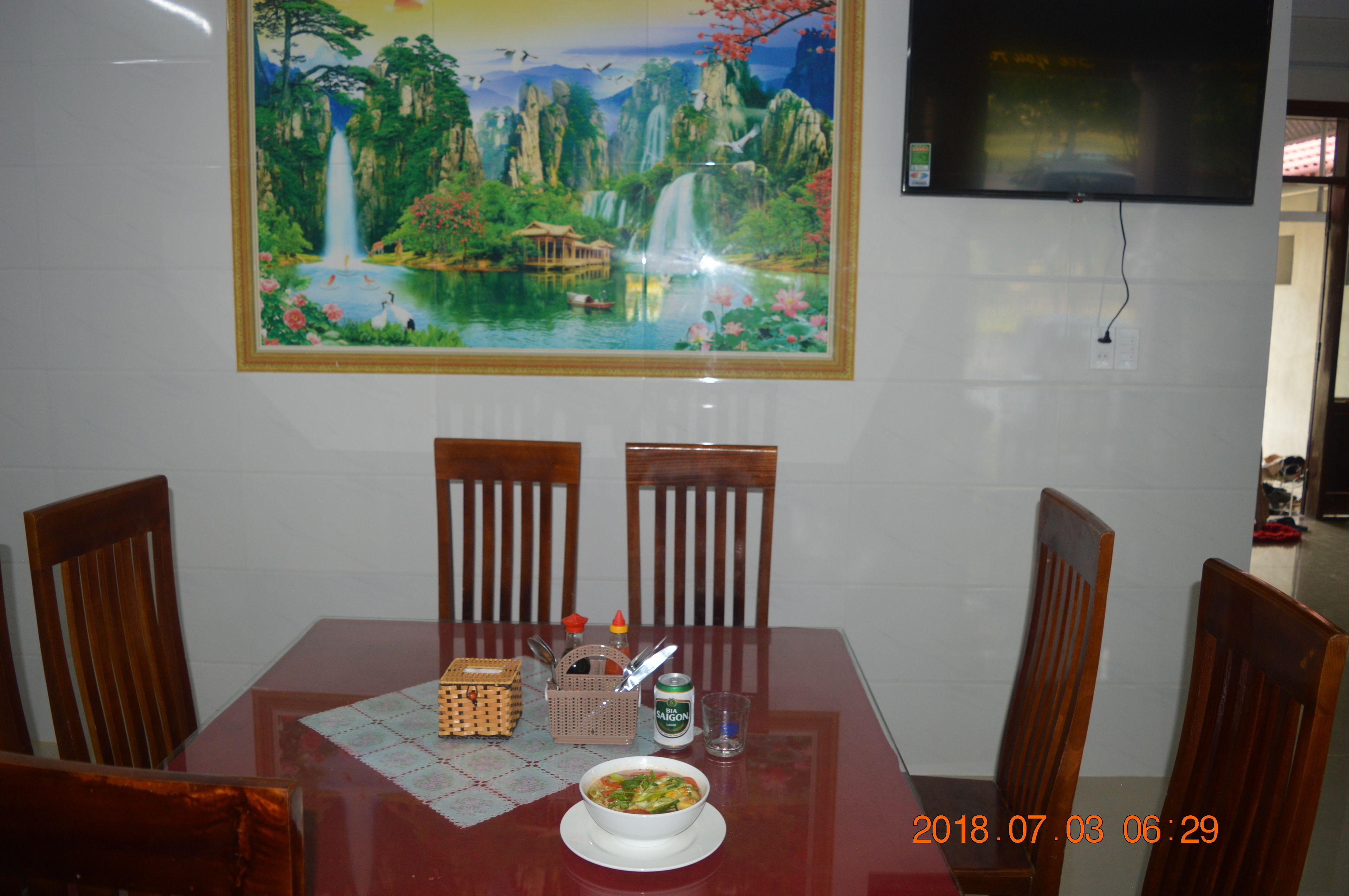 Una mesa de comedor con sillas, un plato de comida, una lata de bebida y una cesta decorativa. Sobre la mesa, un cuadro de un paisaje.