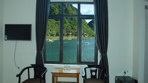 A room with a large window offering a view of a lake with boats and houses. There is a TV on the wall, a small table with glasses, and two chairs. - Son Doong Riverside Homestay (Phong Nha)