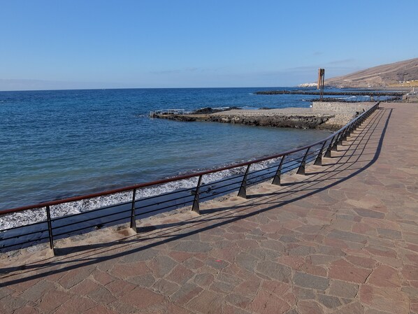 Un paseo marítimo con un camino empedrado de piedra, barandilla metálica y un rompeolas rocoso al fondo.