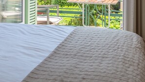 Een bed met een witte en geruite bedspread, een raam met uitzicht op een balkon en groen, en een stoel met een zacht kussen.