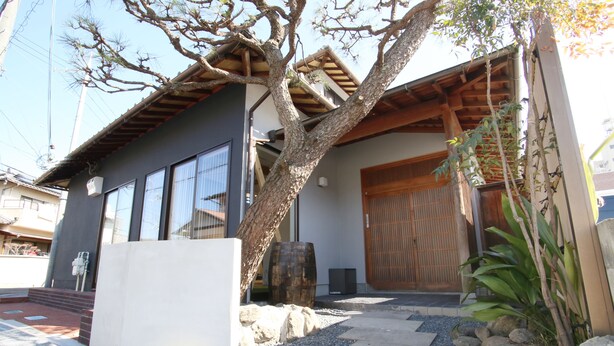 A modern building with a large tree in front, a wooden door, and a stone wall. - BOND - Hostel (Shimamoto)