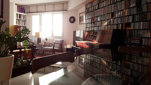 Ein Wohnzimmer mit Klavier, Bücherregal, Pflanze und Fenster.