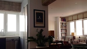 Ein helles Wohnzimmer mit Bücherregal, einem Essbereich mit Stühlen und einer Küche mit Geschirrspüler.