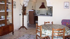 Un comedor con una mesa redonda cubierta con mantel estampado, sillas de madera, un aparador de madera con puertas de cristal, y una cocina visible a través de un arco.