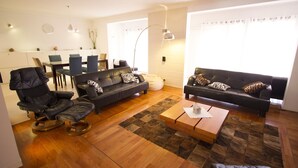 A modern living room with a leather sofa, a black armchair, a wooden coffee table, and a dining area with a table and chairs. - Nightlife but quiet area, modern large home, bayview, garden, bbq, parking (St. George's Bay)