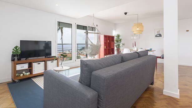 A modern living room with a large grey sofa, a flat-screen TV on a wooden stand, a red bookshelf, and a hanging chair.