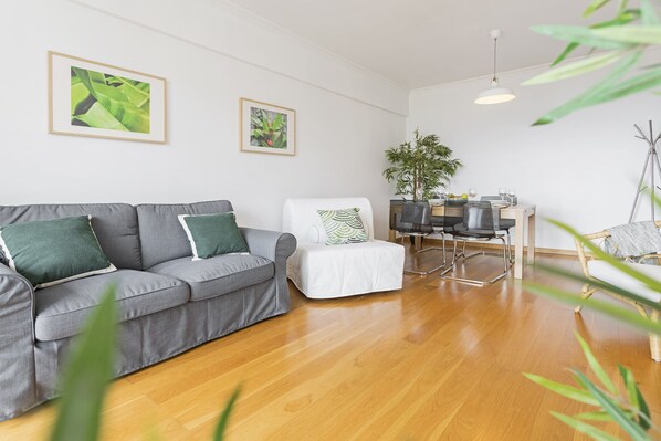 A living room with a grey sofa, a white armchair, a dining table with chairs, and a potted plant.