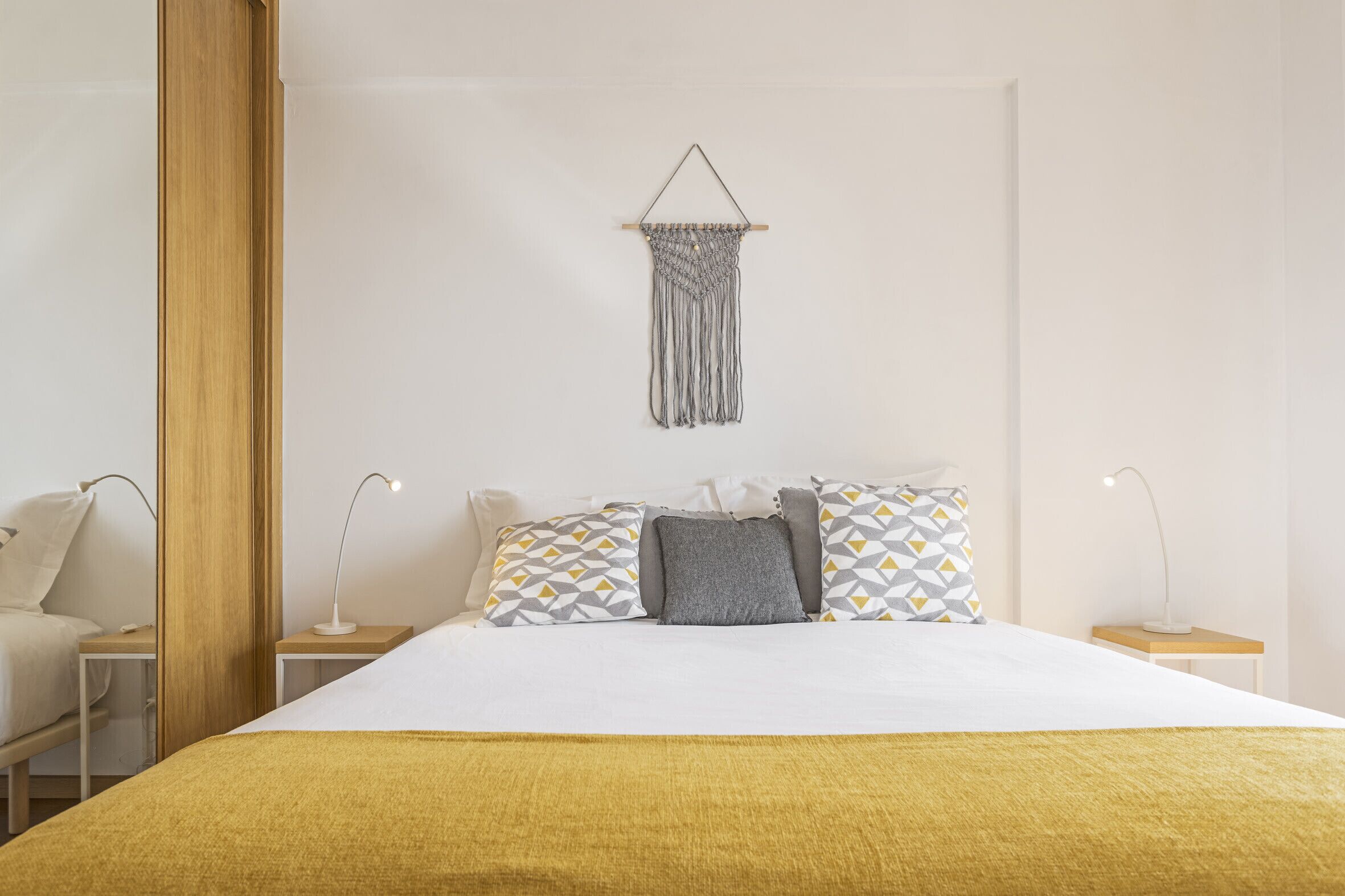 A neatly arranged bedroom with a bed, bedside tables, and a macrame wall hanging.