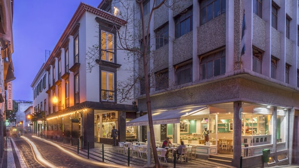 A street view of a building with multiple ground-floor shops and cafes, featuring outdoor seating and a visible restaurant interior. - Downtown Funchal Apartments 2C 25 Fontes. (Funchal)