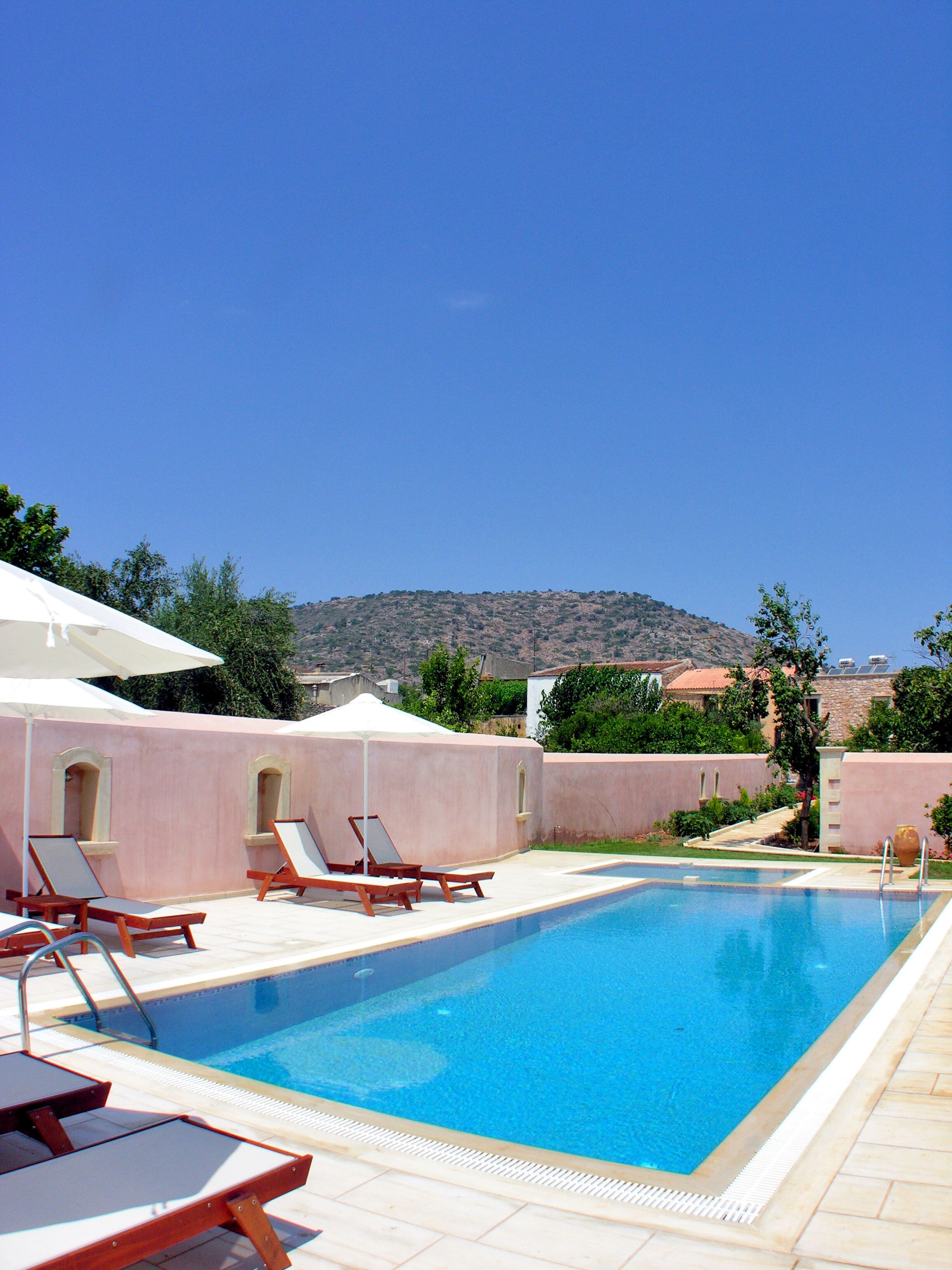 A swimming pool with lounge chairs and umbrellas, surrounded by a pink wall and greenery, with a mountain in the background.