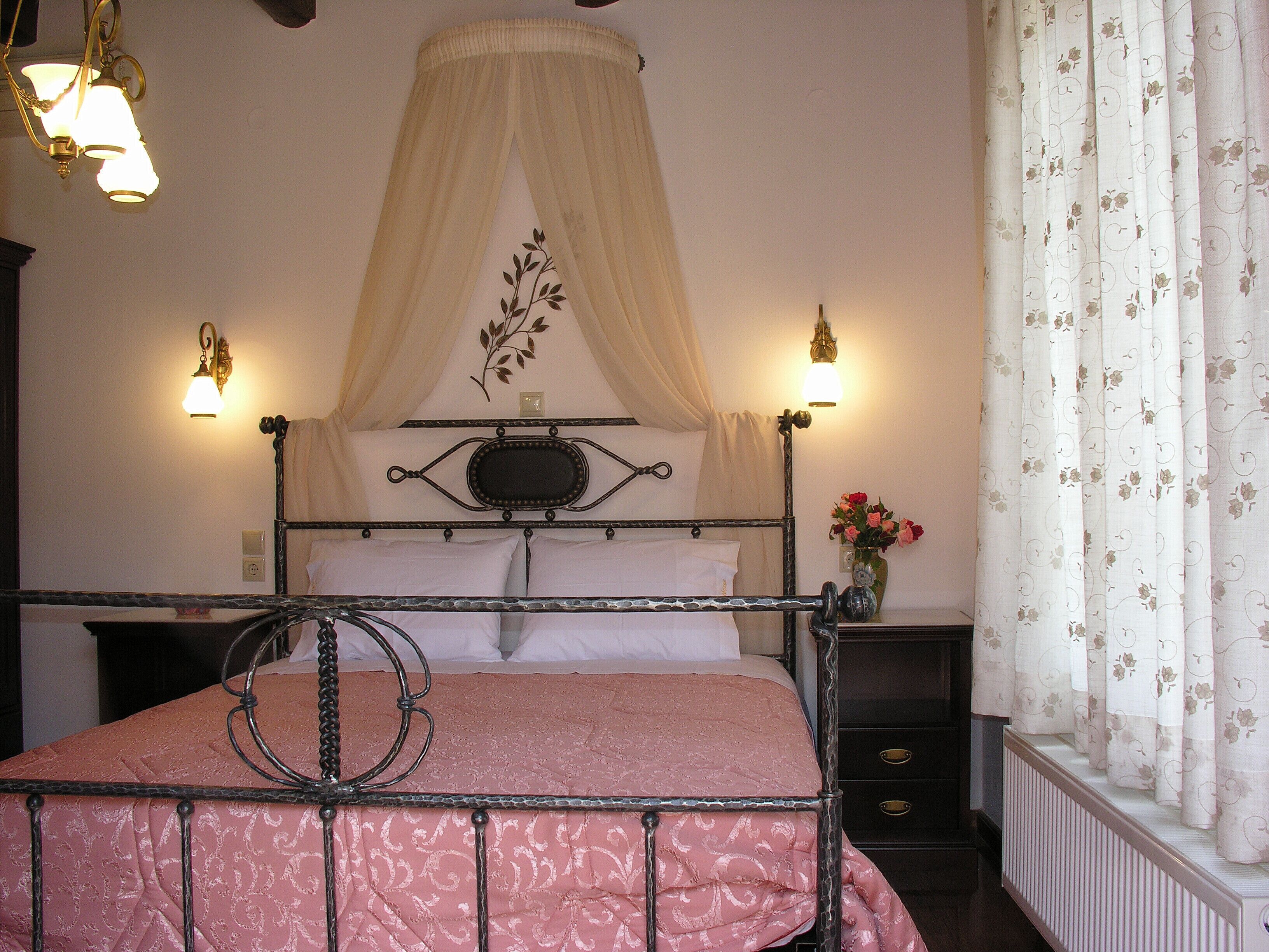 Un lit avec un cadre en métal, un ciel de lit et une couvre-lit rose. Il y a une table de chevet avec un vase de fleurs et une lampe.