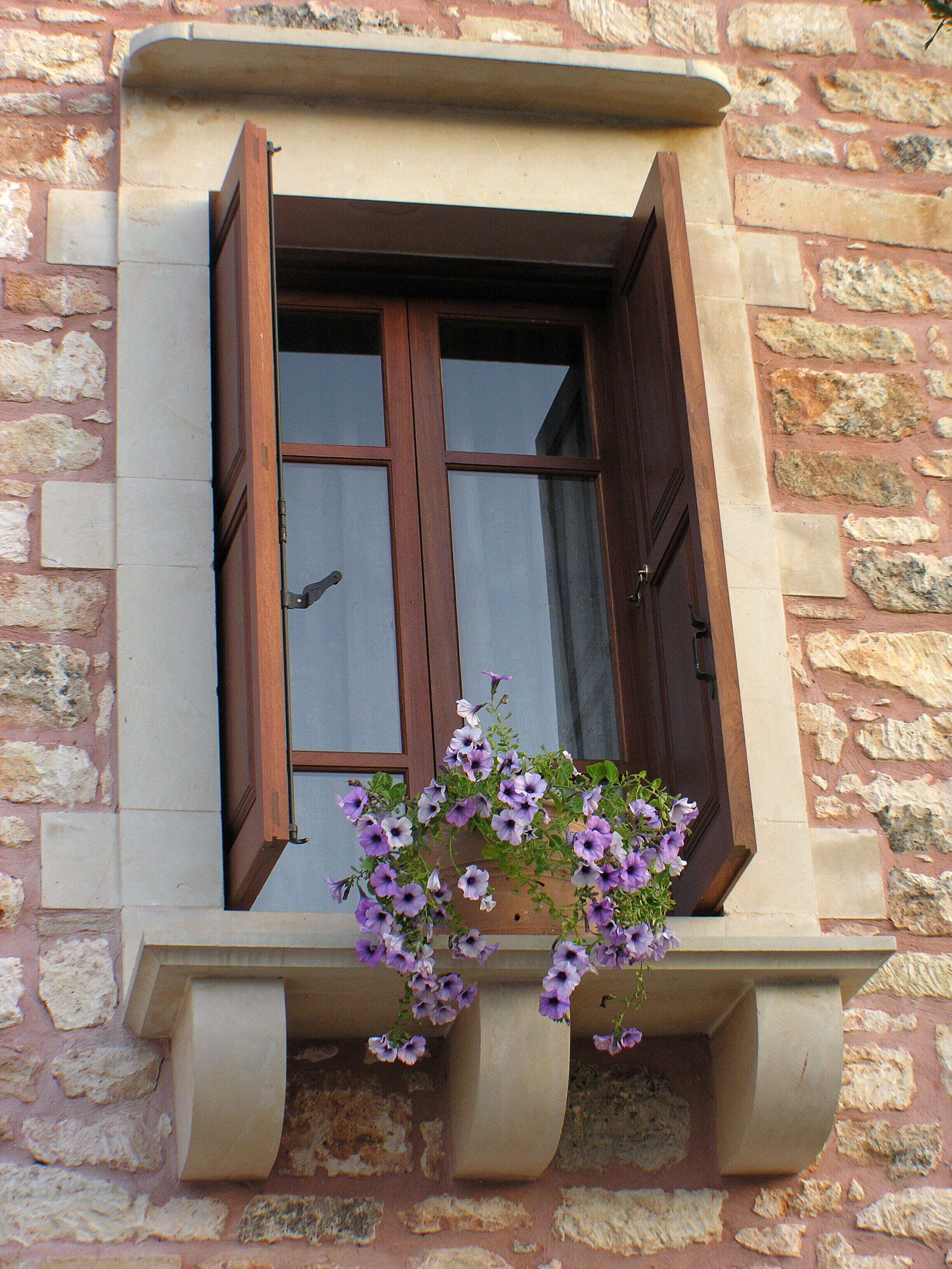Una ventana con contraventanas de madera, un muro de piedra y una maceta colgante con flores moradas.