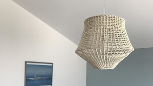 Un dormitorio con una cama blanca, una lámpara colgante tejida, un cuadro con una imagen de un velero y un taburete pequeño de madera.