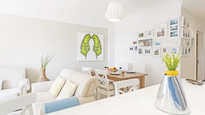 Un salon moderne comprenant un coin repas, des meubles blancs, un vase avec des plantes vertes et un mur décoré de photos encadrées.