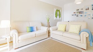 Un salon avec deux canapés de couleur crème, une petite table d’appoint avec une lampe, un vase rempli de fleurs et un mur orné de photos encadrées.