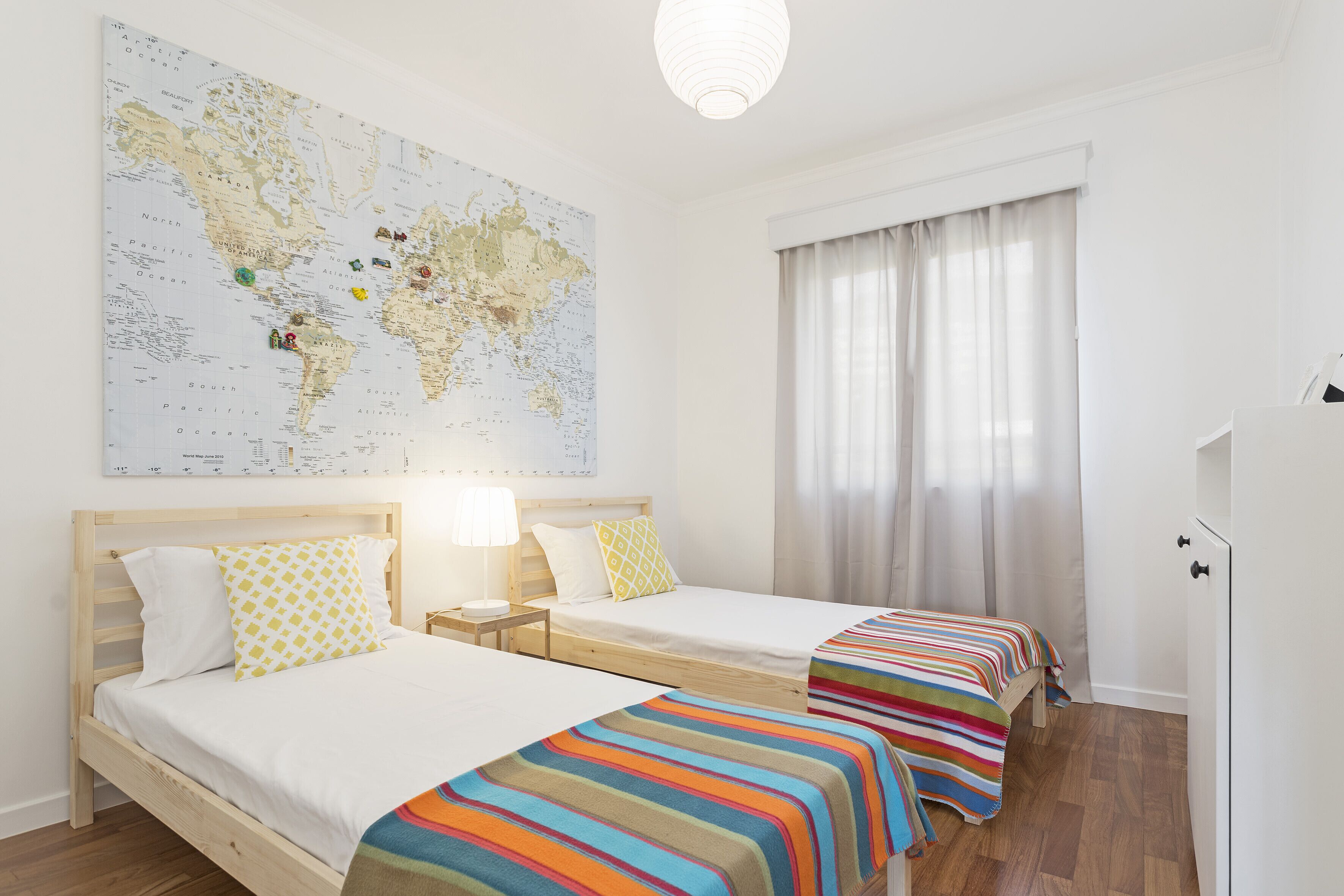 A hotel room with two beds, a large world map on the wall, a window with curtains, and a wardrobe.
