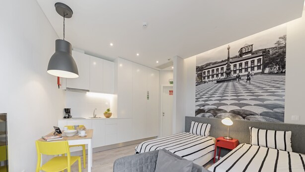 Ein modernes Hotelzimmer mit Essbereich, zwei Betten mit gestreifter Bettwäsche, einem grauen Sofa und einem großen Wandgemälde.