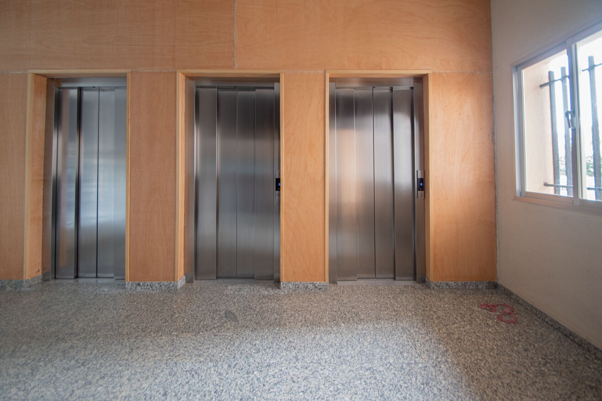 Three elevator doors in a room with a granite floor and a window on the right.