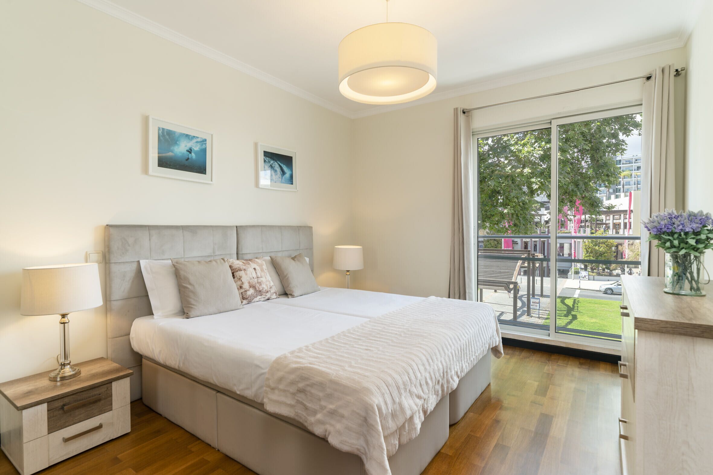 Une chambre avec un grand lit, deux tables de chevet, une commode, une fenêtre donnant sur la ville et un balcon avec une balustrade.