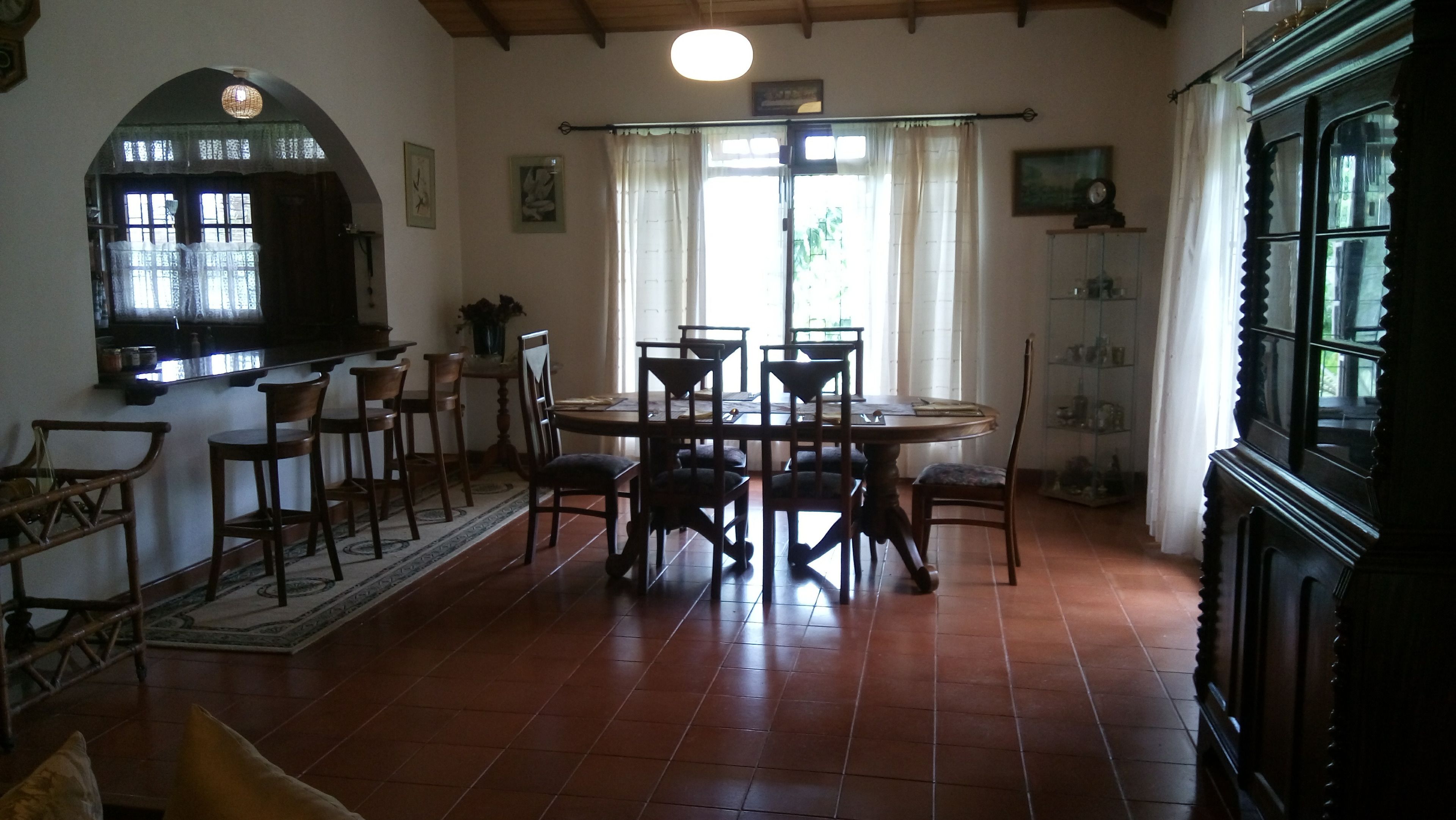 A dining area with a round table and multiple chairs, a kitchen area with a counter and chairs, and a large window with curtains.