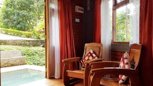 A wooden chair with a cushion, a window with red curtains, and a view of a garden and pool. - Tea Leaf Retreat Peradeniya (Doluwa)