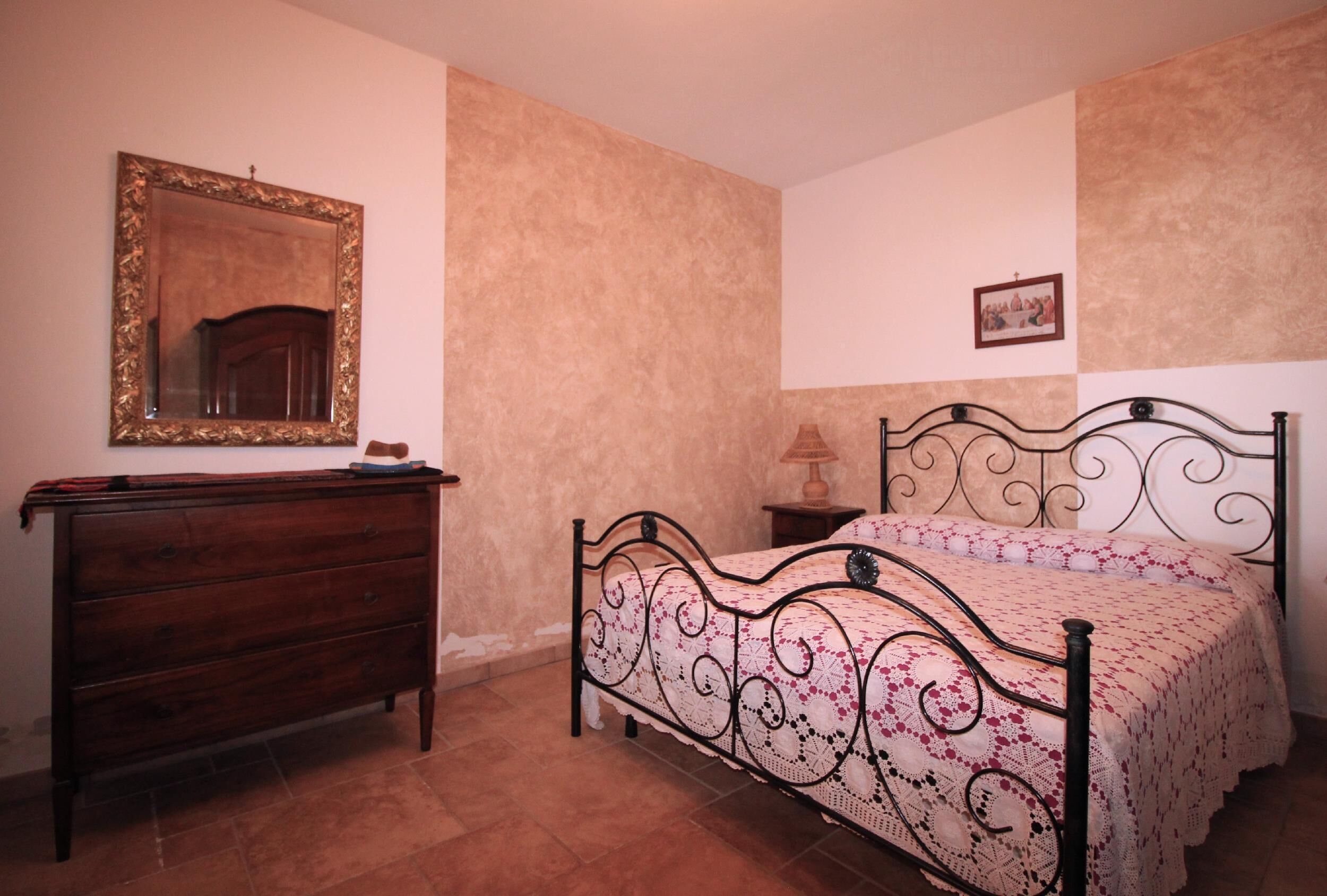 Een slaapkamer met een houten dressoir, een bed met metalen frame, een spiegel en een kleine tafel met een lamp.
