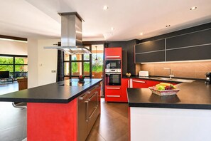 A modern kitchen with a central island, red cabinetry, and a large window overlooking greenery.